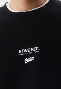 Černá mikina s bílým kulatým výstřihem. Obsahuje tištěné logo a text: "STWD REC. 0000 P & B 0091 EST 091" v městském písmu.