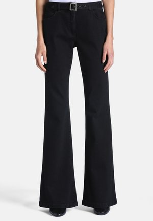Personne portant un pantalon noir évasé taille haute avec une ceinture à boucle en strass et des chaussures noires, debout devant un fond clair uni.