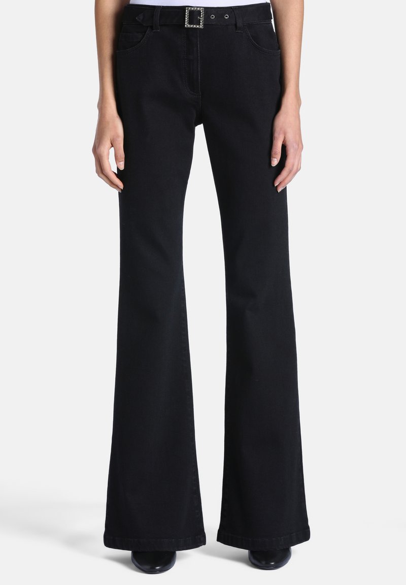 Personne portant un pantalon noir évasé taille haute avec une ceinture à boucle en strass et des chaussures noires, debout devant un fond clair uni.