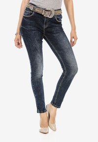 Dunkelblaue Jeans mit einem verblassten Muster, ausgestattet mit einem geschmückten Gürtel, schmal geschnitten und mit mittelhoher Taille, kombiniert mit beigen Absätzen.