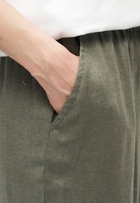 Χέρι που έχει βυθιστεί στην τσέπη παντελονιού από ελιά πράσινο ύφασμα, συνδυασμένο με λευκό τοπ που φαίνεται στη μέση.