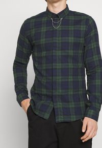 Camisa de cuadros verde y azul marino de manga larga con un cuello estrecho y un detalle de cadena en la parte frontal. Fabricada en tejido tejido con una textura suave.