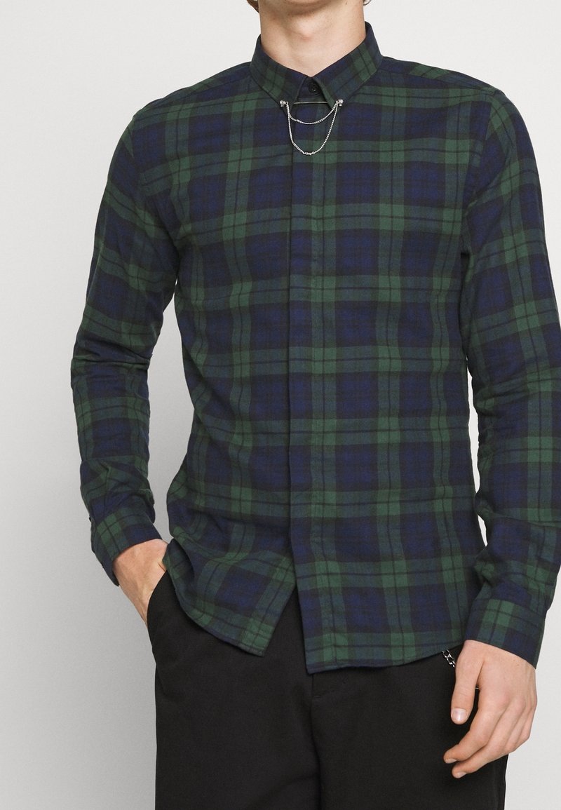 Camisa de cuadros verde y azul marino de manga larga con un cuello estrecho y un detalle de cadena en la parte frontal. Fabricada en tejido tejido con una textura suave.