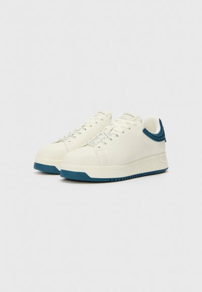 Des baskets blanches avec un dessus lisse, des accents bleus sur le talon et la semelle, et des lacets plats. Détails textuels sur le côté et une semelle rembourrée.