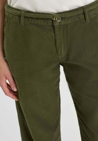 La mano de una mujer reposando cerca del bolsillo de unos pantalones de pana verde oliva con un botón visible y una hebilla de doble anilla en D.