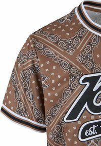 Brūns džersijs ar paisley rakstu un baltām akcents. Iezīmēts ar apaļu kakla izgriezumu, īsām piedurknēm un lieliem melniem burtiem ar logotipu.