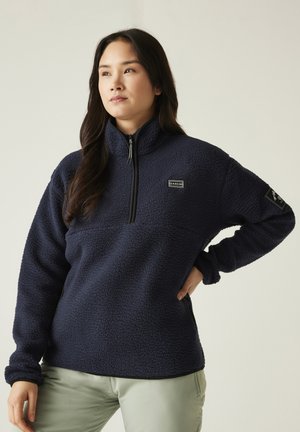 Junge Frau mit langen dunklen Haaren trägt einen marineblauen Fleecepullover und helle Hosen, steht mit einer Hand in der Hüfte.