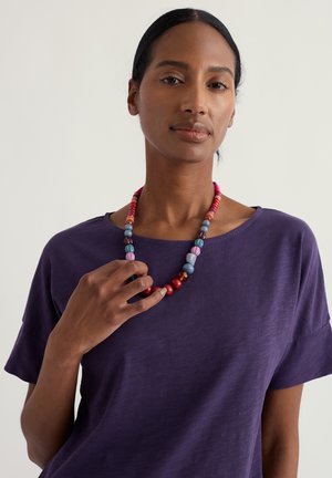 Collier coloré en perles comprenant des perles rouges, roses, bleues et violettes de différentes formes. Le modèle porte une chemise violette à manches courtes.