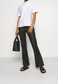 Persona che indossa una camicia bianca a maniche corte, pantaloni in pelle neri a campana, sandali neri e tiene in mano una borsa a secchiello nera.