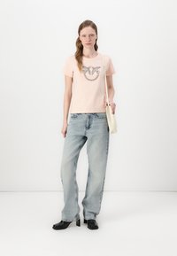 Lys rosa tettsittende T-skjorte med metalliske dekorative elementer i et sirkulært mønster, kombinert med lyseblå baggy jeans og svarte høyhælte støvler.