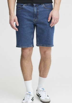 Man in blauwe spijkershorts, witte sportsokken en witte Adidas-sneakers met zwarte strepen, staand tegen een effen achtergrond.