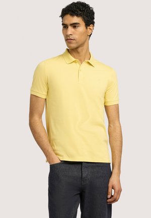 Mann trägt gelbes kurzärmeliges Polohemd und dunkle Hose, steht mit einer Hand in der Tasche und schaut leicht zur Seite.