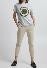 Grijze t-shirt met een rond groen glimlachend gezicht grafiek en tekst "MINDER PERFECTIE" in het zwart, gecombineerd met beige strakke broek en zwarte instapschoenen.
