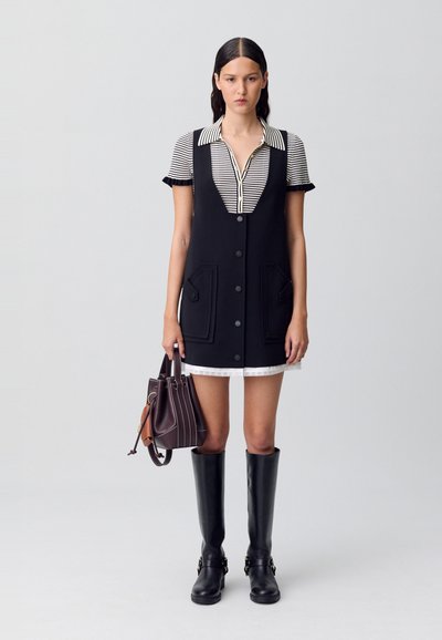 Donna che indossa un vestito smanicato nero con bottoni sopra una camicia a righe a maniche corte, tiene una borsa marrone e indossa stivali neri al ginocchio.