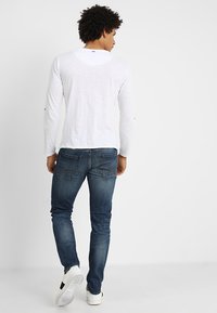 Langärmliges weißes Shirt mit Rundhalsausschnitt, kombiniert mit blauen Slim-Fit-Jeans und weißen Sneakers. Zeigt einen lässigen, figurbetonten Stil.