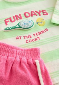 T-shirt a righe verde chiaro e bianco con ricami rosa, una toppa a forma di racchetta da tennis e una grafica di una pallina da tennis gialla. Pantaloni corti in terry rosa.