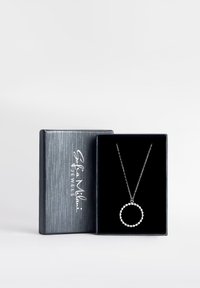 Silverhalsband med en cirkulär hänge som har en pärldesign, visad i en låda klädd med svart sammet och med en metallgrå utsida.