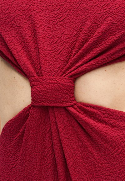 Κοντινό πλάνο φορέματος από κόκκινο υφασμάτινο ύφασμα με λεπτομέρεια δέσιμου κόμπου που αποκαλύπτει μερικό δέρμα από κάτω.