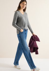 Grauer V-Ausschnitt-Pullover, blaue gerade Jeans, weiße Sneakers mit burgunderfarbenen Akzenten, hält einen burgunderfarbenen Hoodie. Glatte Texturen, lässiges Design.