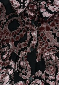 Chemise à texture velours avec un motif floral rose poussiéreux et rouge foncé sur fond noir, dotée d’un col et de boutons-pression noirs.