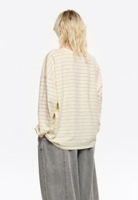 Camicia a maniche lunghe a righe di colore giallo chiaro con sottili strisce scure. Tessuto morbido, vestibilità ampia e scollatura rotonda. Abbinata a pantaloni grigi larghi.
