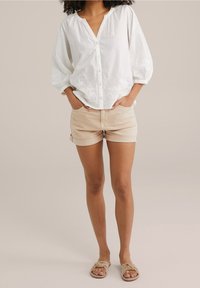 Blusa blanca de botones con mangas abullonadas y detalles bordados, combinada con pantalones cortos beige enrollados y sandalias planas marrones.