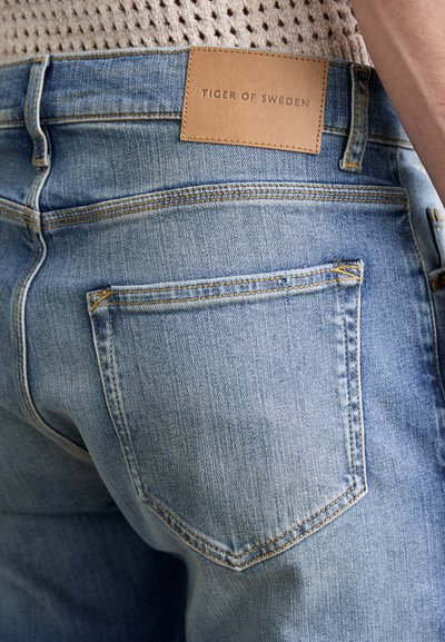 Svetlo modri jeans iz denima z klasičnim dizajnom, s svetlo rjavo usnjeno nalepko z vtisnjenim napisom "TIGER OF SWEDEN" in podrobnostmi šivanja na zadnjem žepu.