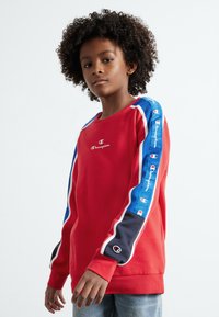 Sudadera roja con rayas de acento en azul y negro en las mangas. Presenta un pequeño logo blanco en el pecho y puños acanalados. Textura de tela suave.