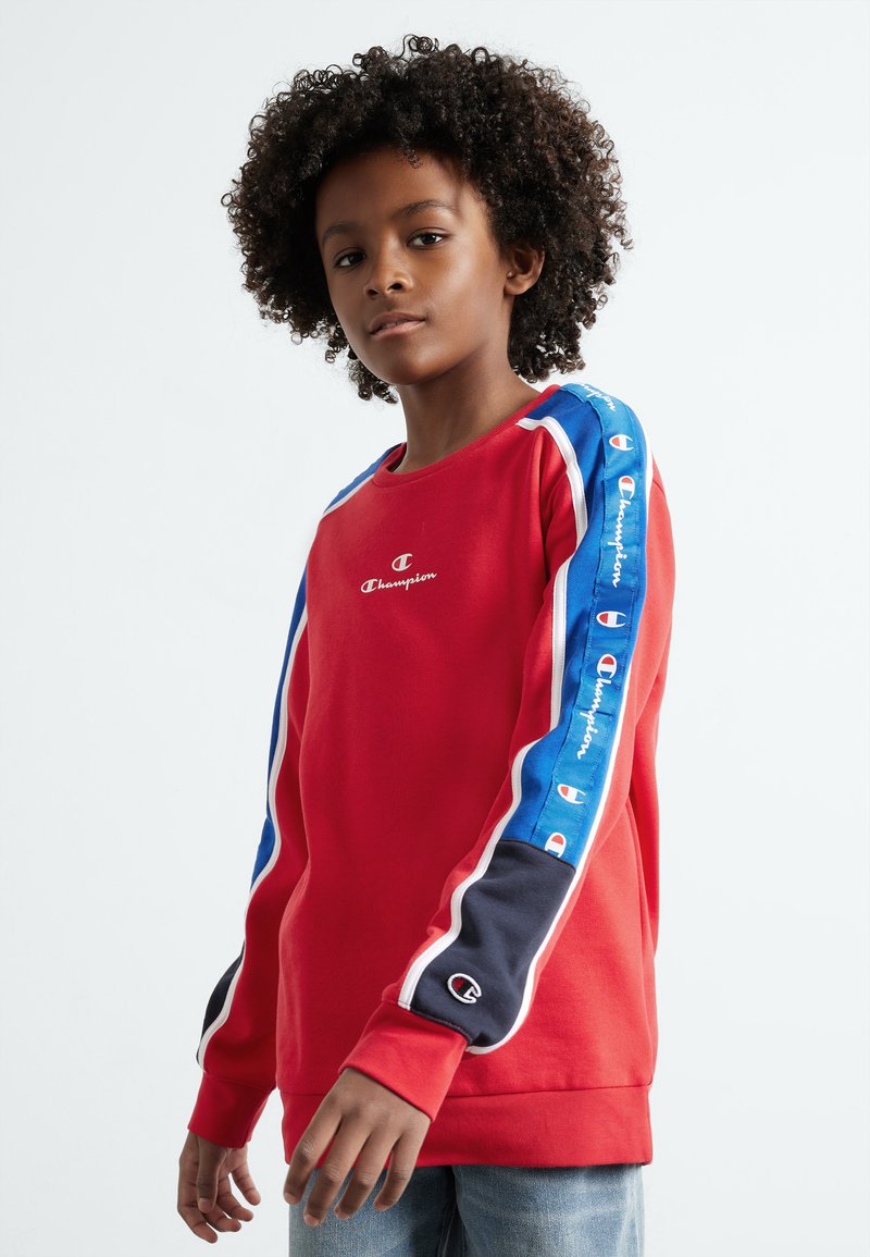 Sudadera roja con rayas de acento en azul y negro en las mangas. Presenta un pequeño logo blanco en el pecho y puños acanalados. Textura de tela suave.