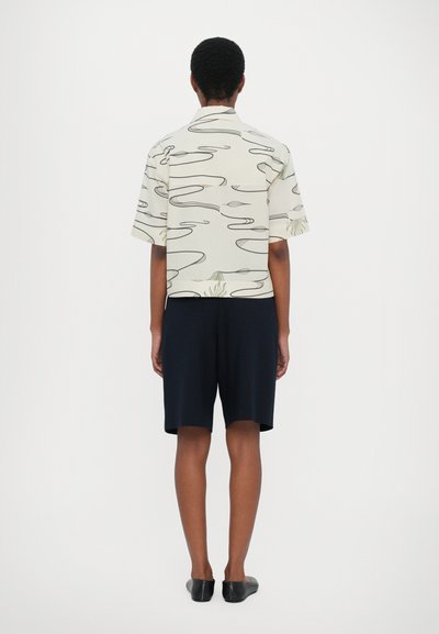 Camicia a maniche corte con motivo a linee ondulate in beige e verde, abbinata a pantaloni corti blu scuro e ballerine nere. Solo vista posteriore.