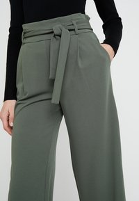 Zelené široké kalhoty vyrobené z hladkého materiálu, s vysokým pasem a detailem na zavazování, bočními kapsami a přizpůsobeným střihem.