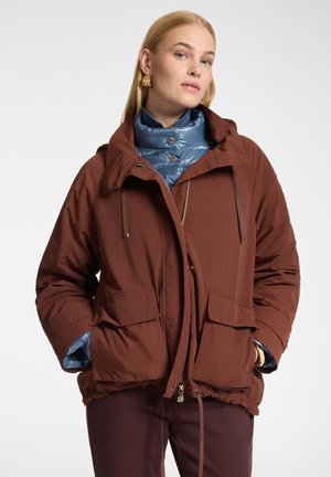 Donna che indossa una giacca imbottita marrone sopra un top trapuntato blu con zip, con le mani nelle tasche della giacca e orecchini d'oro, su uno sfondo semplice.