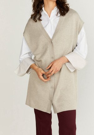 Beige sleeveless cardigan with a V-neck, button closure, and textured knit fabric. Worn over a white shirt with wide cuffs and burgundy pants.