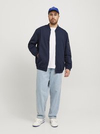 Granatowa kurtka bombowca, biała koszulka, jasnoniebieskie luźne dżinsy i białe sneakersy z niebieskimi akcentami. Model nosi niebieską czapkę.