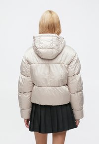 Lyshåret person set bagfra iført en beige dunjakke med hætte og et sort plisseret skørt.