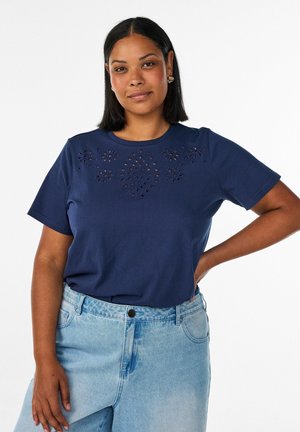 Donna con carnagione media che indossa una maglietta blu navy a maniche corte con motivo floreale traforato e jeans azzurri a vita alta, mano sul fianco.