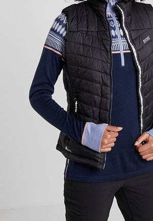 Persoon draagt een zwart doorgestikt vest over een marineblauwe, patroonrijke trui met rits en duimgaten, en houdt de rits van het vest dicht bij de borst vast.