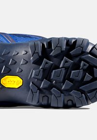 Blå trail-sko sula med en robust textur, djupa multidirektionella klackar för grepp och en gul Vibram-logotyp i mitten.
