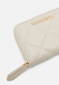 Valentino quiltet læderpung i lys beige med guldlogo og lynlåslukning, med fine syninger og en tekstureret overflade.