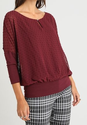 Frau trägt eine durchsichtige, bordeauxrote, strukturierte Bluse mit kleinem Knopf-Schlüsselloch und schwarz-weiß karierten Hosen.