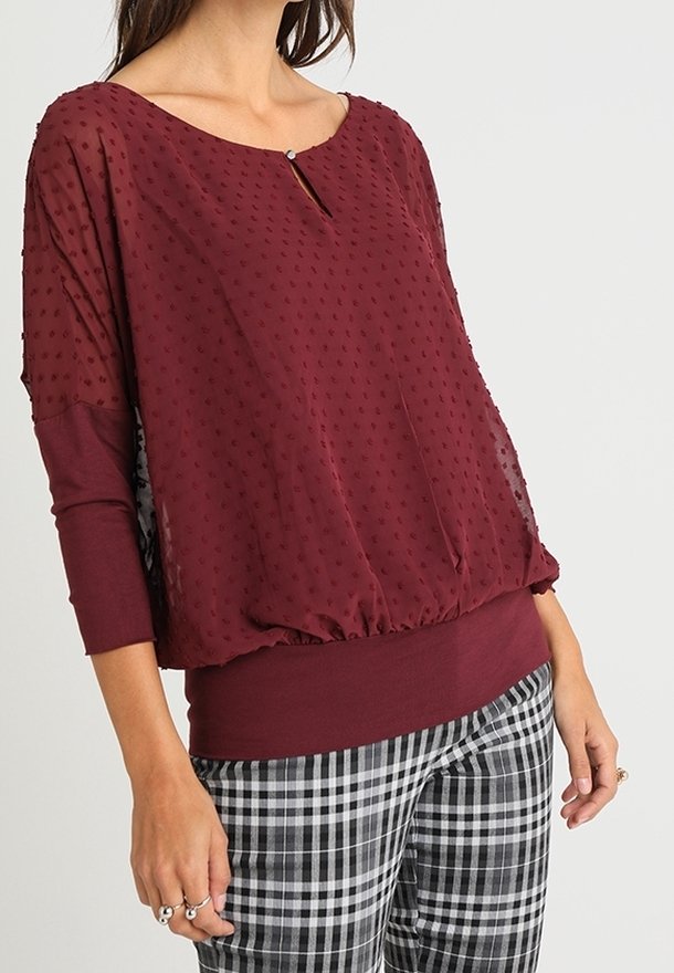 Femme portant un chemisier transparent texturé bordeaux avec un petit keyhole boutonné et un pantalon à carreaux noir et blanc.
