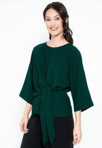 Blusa verde con maniche ampie, caratterizzata da un nodo in vita e una superficie liscia. Collo tondo semplice con dettagli di pieghe sottili.