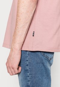 Camicetta corta a maniche corte di colore rosa chiaro con orlo curvo, abbinata a jeans in denim blu. Presenta una piccola etichetta nera del marchio sul lato della camicetta.