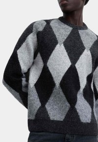 Persona che indossa un morbido maglione argyle nero e grigio con grandi motivi a diamante, abbinato a pantaloni scuri, su uno sfondo semplice.