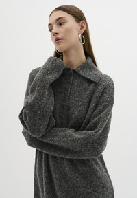 Maglione grigio in lana con colletto, maniche lunghe e chiusura frontale con bottoni. Tessuto morbido e texture. Modella in posa con le braccia incrociate.