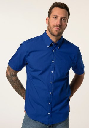 Uomo con capelli corti e barba che indossa una camicia azzurra a maniche corte con bottoni, posando con le mani dietro la schiena.