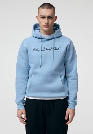 Młody mężczyzna w jasnoniebieskiej bluzie z kapturem z napisem „Diverse Sport Club”, stojący na jasnym, jednolitym tle.