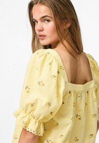 Blusa gialla con maniche corte a sbuffo, schiena quadrata, tessuto testurizzato e motivo floreale che presenta piccoli fiori gialli e accenti verdi.