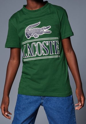 Camiseta de algodón verde con un gran gráfico de un cocodrilo y el texto "LACOSTE" en blanco y azul marino. Mangas cortas y cuello redondo.