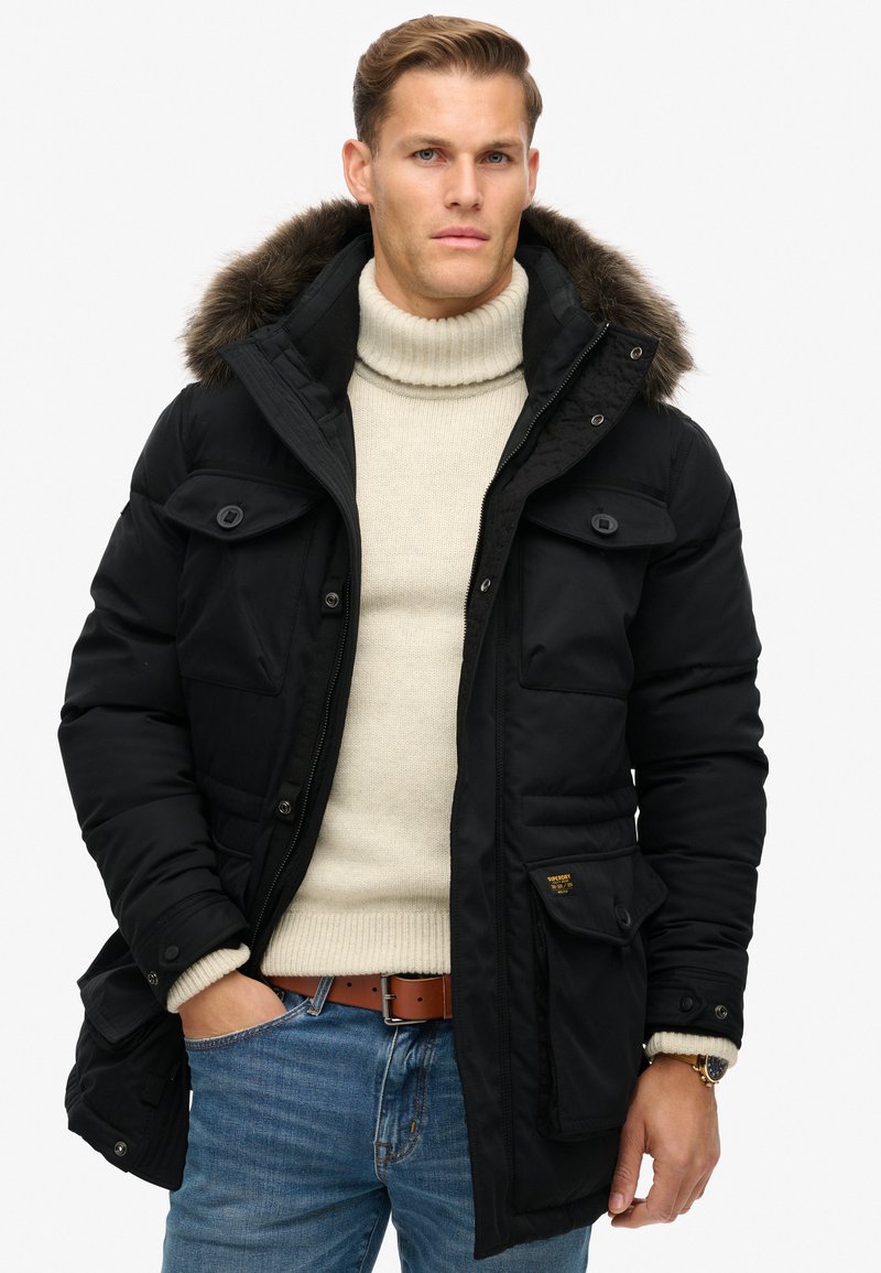 Zwarte pufferjack met een bontvoering in de kap, meerdere zakken en een strak ontwerp. Gekocht over een beige turtleneck, gecombineerd met blauwe jeans.